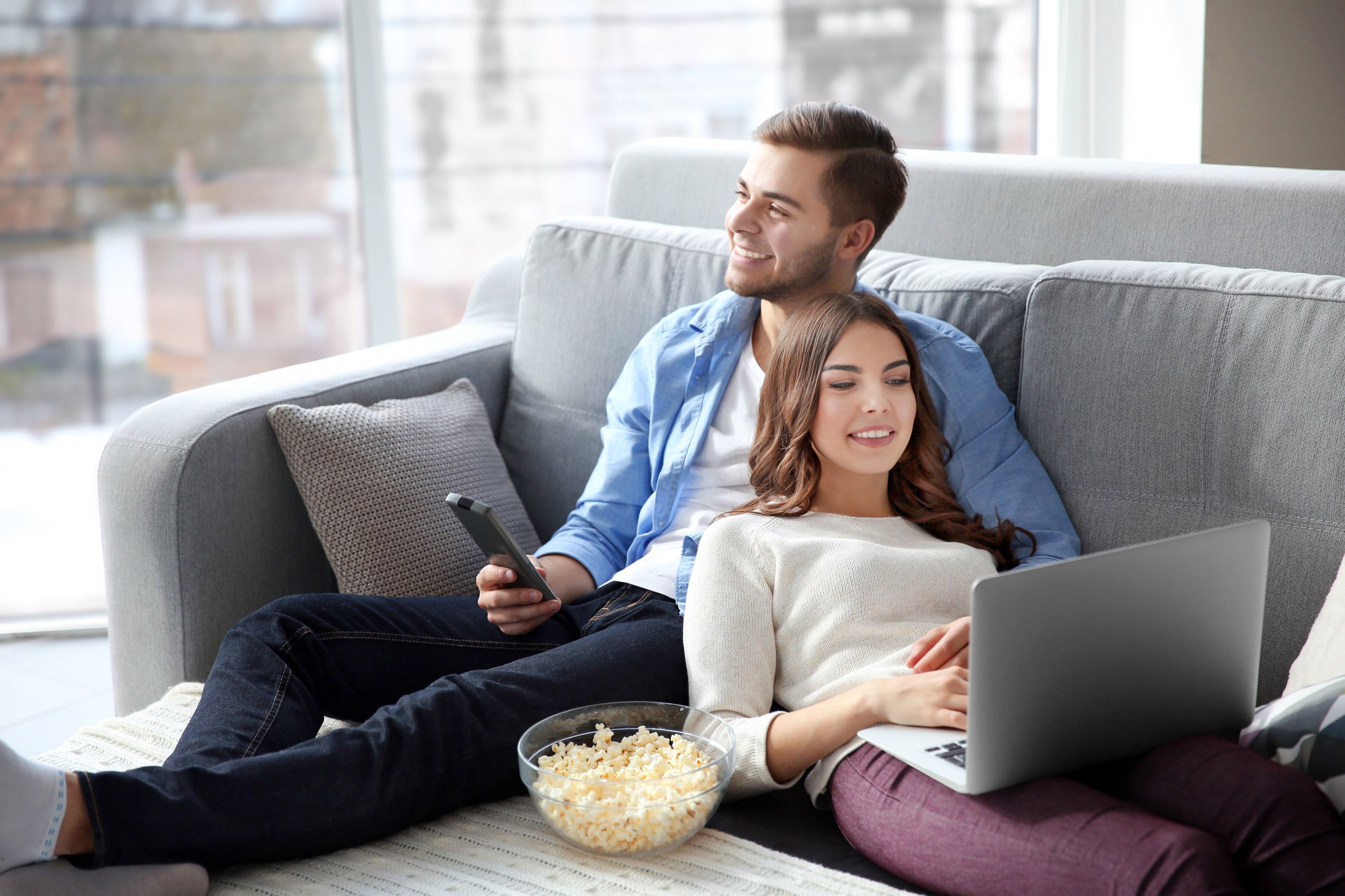 Young man watching TV and woman using laptop on a sofa at home